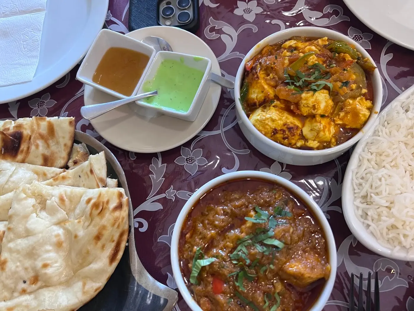 Curries en cuencos con arroz basmati, naan y dos salsas, vistos desde arriba, sobre un mantel burdeos