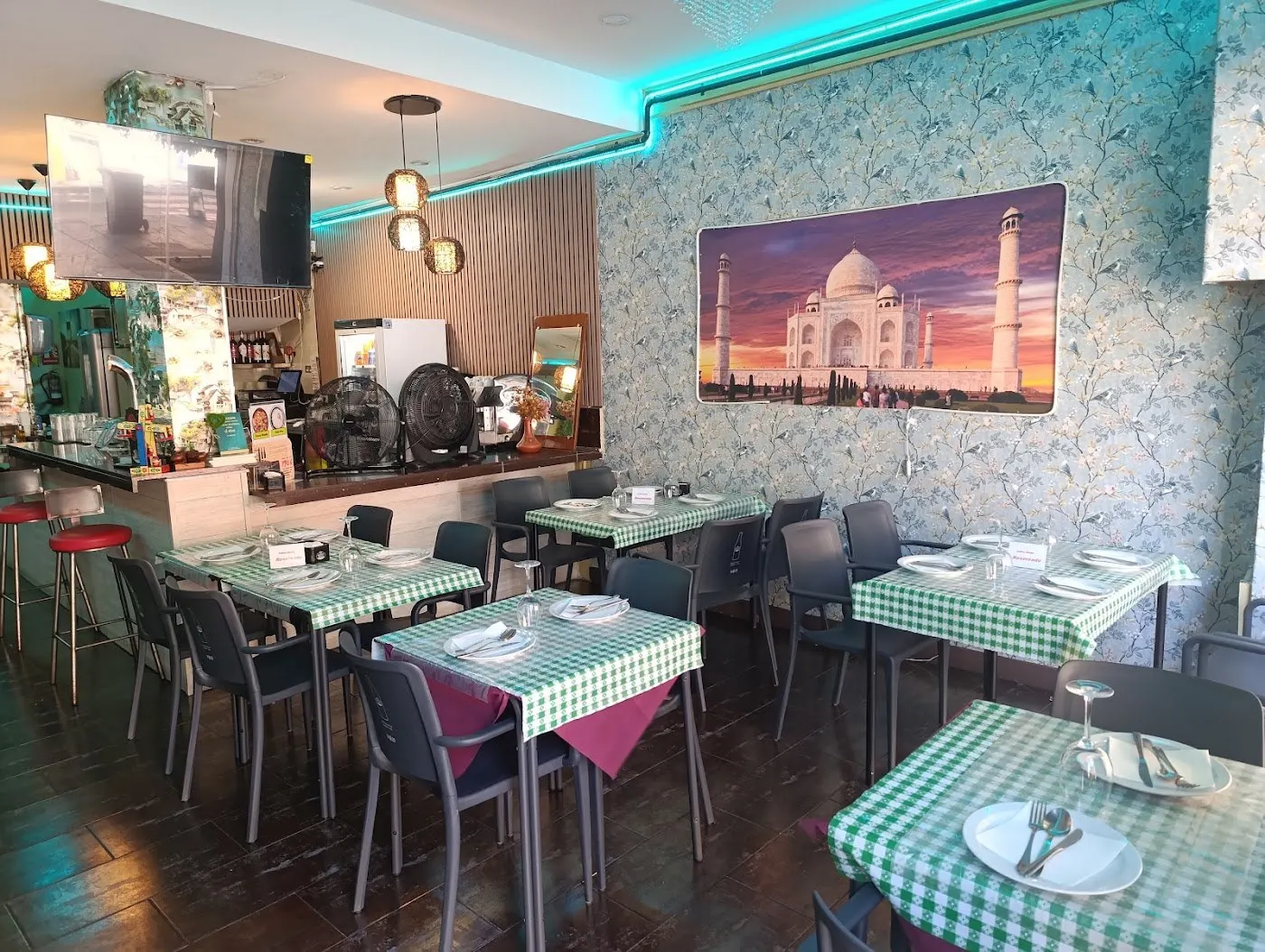 Comedor con mesas de cuadros verdes, una barra al fondo y una fotografía del Taj Mahal en la pared
